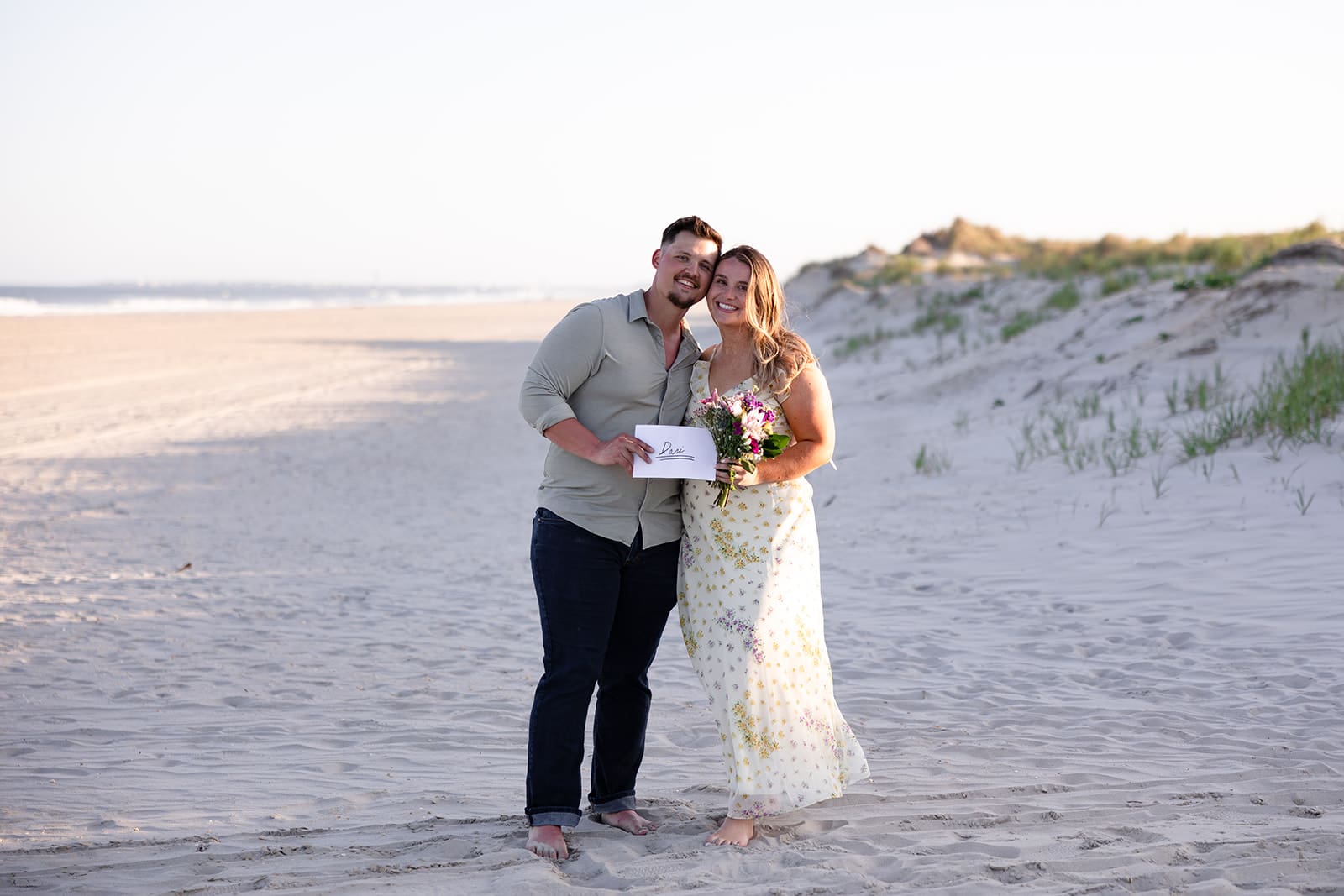 Philadephia PA photographer captures a surprise beach proposal in Sea Isle City NJ. The newly engaged couple embraces with the golden hour sun casting a warm glow on the sand dunes. They are looking at the camera and holding a letter and flowers.